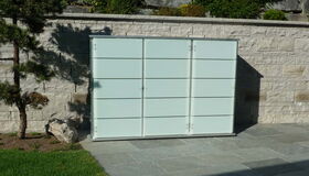 . The image shows a modern outdoor setting with a sleek storage unit or cabinet against a stone wall. The unit has frosted glass panels, and the area is landscaped with a tree and some greenery. The ground is covered with flat stone tiles, and there is a grassy area nearby.