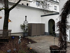 . Ein Garten mit Sitzbank, Vogelhaus, Skulptur, Pflanzengitter und Schrank vor einem weißen Gebäude.