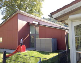 . Ein modernes Gebäude mit roter Fassade, kleinem Rutschspielplatz, und einer Person im Freien.