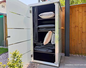 Ein geöffneter Gartenschrank mit Kissen und Decken auf Regalen, umgeben von einem Holzzaun und Steingarten.