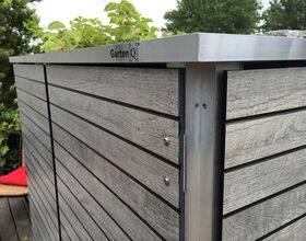 . The image depicts a wooden storage unit with horizontal slats and a metal frame. The structure is marked with "Garten[Q]," indicating a brand or model. It is located on a deck and surrounded by greenery.