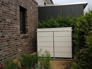 . Ein moderner, weißer Gartencontainer neben einer Backsteinmauer, umgeben von Pflanzen und einem Kiesweg.