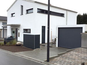 . The image shows a modern, minimalistic house with a white exterior. It features large windows and a flat roof. There's a driveway leading to a gray garage door and a small front yard with landscaping, including gravel and a few plants. A streetlight is visible in front of the house. The design is clean and contemporary.