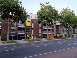 . The image shows a street with a modern residential building in the background. The building has a brick facade with large windows and some colorful accents on the lower level. There are several trees lining the street in front of the building, and a few people can be seen walking or standing near the entrance. The street in the foreground has marked lanes and sidewalks.