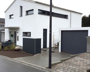 . The image shows a modern, two-story house with a minimalist design. It has a white façade with dark-framed windows and a flat roof. In front of the house, there is a pathway and a yard area with plants and a large rock. The house features a garage with a dark-colored door. A streetlight is visible on the sidewalk. The overall setting appears to be in a suburban residential area.