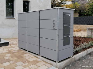 . The image shows a modern outdoor parcel locker system. It is installed on a paved area outside a building. The locker system is gray and has several compartments for storing parcels, with a sleek and minimalist design. The word "PAKETE" is printed on the side. The surrounding area includes some greenery and paved stones.