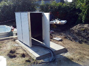 . The image shows an outdoor scene where two people are working. There is a small, open structure that resembles a utility or storage cabinet. It is situated on a concrete slab with cables or hoses leading into it. Nearby, there are building materials and tools on a table, indicating some construction or installation activity. The area is surrounded by grass and trees.