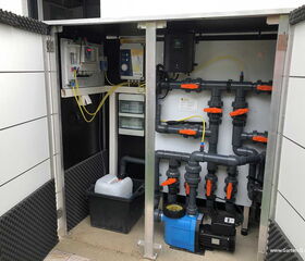 . The image shows a technical cabinet or enclosure containing a pool or water treatment system. It includes:

1. **Pipes and Valves**: Several grey PVC pipes with orange-handled valves for directing water flow.

2. **Pump**: A blue water pump located at the bottom of the cabinet.

3. **Control Panel**: A control panel on the left with cables and a display for monitoring and controlling the system.

4. **Chemical Container**: A white container, likely for storing chemicals or treatment solutions.

5. **Electrical Components**: Wiring and electronic components for system operation.

This setup is often used for pool or water filtration systems.