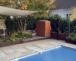 The image depicts an outdoor scene featuring a covered swimming pool surrounded by a spacious patio area. There is a garden with various plants and a tree with colorful autumn foliage. A modern outdoor shower and a shade sail are also part of the setup, with a house visible in the background.
