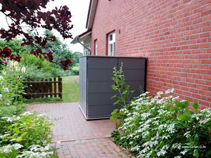 . The image depicts an outdoor setting with a brick pathway leading to a wooden fence. There is a red brick building to the right, and adjacent to it, a modern, gray storage unit. The foreground has various plants, including flowers and leafy shrubs. A tree with red leaves partially frames the top left corner.