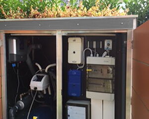 The image shows a cabinet housing various pool equipment. It includes different pumps, filters, and control devices typically used for maintaining a swimming pool's water quality and circulation. The setup appears organized and is located outdoors, with a leafy background.