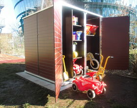 . The image shows an open storage shed in a garden or outdoor setting. Inside the shed, there are several items:

1. Shelving with various toys and equipment
2. Red and yellow toy vehicles 
3. Buckets and small pails stacked on the shelves
4. A small yellow shovel leaning against the side
5. A balance bike or similar ride-on toy

The background includes some modern buildings and greenery.