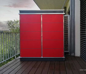 Rote Gartenbox auf einem Balkon neben Glasfront und Geländer mit Ausblick auf Bäume.