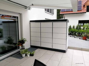 . Großer moderner weißer Schrank auf Terrasse, umgeben von Pflanzen und Sonnenschutz, Haus mit Solarmodulen im Hintergrund.