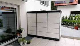 . Großer moderner weißer Schrank auf Terrasse, umgeben von Pflanzen und Sonnenschutz, Haus mit Solarmodulen im Hintergrund.