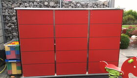 . The image depicts a backyard or garden area with several objects:

1. A large red cabinet or storage unit with metal trim and horizontal lines, likely for outdoor storage.
2. A transparent plastic box containing colorful blocks or toys, possibly for children.
3. A metal wire mesh filled with rocks, functioning as a gabion wall or fence.
4. A child’s spring rider playground toy with a seat shaped like a red animal, on a green spring.
5. Part of a garden in the background with plants and a pathway.

The setting appears to be a modern and organized outdoor area, possibly for a residential space.