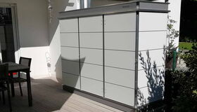 . Ein Gartenschrank auf einer Terrasse neben einem Tisch mit Stühlen, umgeben von Pflanzen und Sonnenschein.