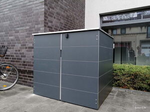 . Ein grauer Metallschuppen neben einem Fahrrad und einer Hecke, vor einem Ziegelgebäude mit Fenster.