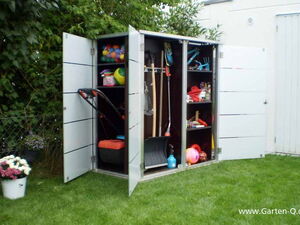 . The image shows a garden storage cabinet with open doors, revealing various items inside. In the left section, there's a lawn mower. The middle section contains gardening tools like rakes and spades, a snow shovel, and a blue spray bottle. The right section has shelves filled with colorful balls, a basket, a bucket, cans, and other small gardening or household supplies. There's also a fence with plants in the background and some flowers in a pot on the grass.