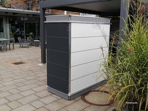 . Ein moderner Müllcontainer auf einer gepflasterten Fläche neben einem Café mit Stühlen und Pflanzen.