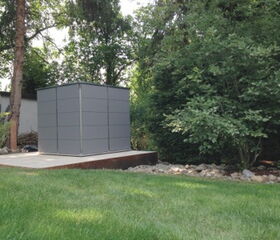 . This image shows a small structure, possibly a garden shed, situated in a backyard setting. The shed has a modern, sleek design with gray panels and is placed on a concrete slab. Surrounding the shed are trees and bushes, with a well-maintained grassy lawn in the foreground.