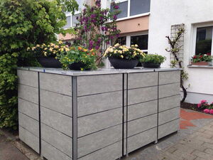. Große, graue Recycling-Tonnenbox mit blühenden Pflanzen darauf, neben einem Haus und grünem Laub.