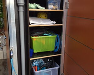 The image shows an outdoor storage cabinet with several shelves. Inside, there are:

1. A vertical pipe along the left side.
2. Various items on the top shelf, including a transparent box.
3. A yellow ball and what looks like a folded towel or cloth on the second shelf.
4. A green plastic crate with more items inside and a hose or cable on the third shelf.
5. A transparent storage bin with assorted items on the bottom shelf.

The cabinet door is open, and there's a blue rope or cord attached to the door handle.
