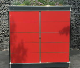 . The image shows a modern red storage unit, likely made for outdoor use. It is placed on a tiled surface with grass in the foreground. Behind it, there's a wall made of wire cages filled with rocks. The roof of the storage unit has greenery or plants growing on it. The setting appears to be an outdoor garden or patio area.