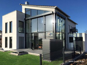 . The image shows a modern building with a sleek, minimalist design. The structure features large glass windows and a combination of concrete and metal elements, providing a contemporary aesthetic. The building appears to be a house situated in a yard with a small lawn and paved area. There's also a metal enclosure or box in the foreground near a fence.