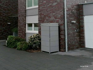 . Ein grauer Metallschrank steht neben einem Ziegelgebäude, umgeben von Sträuchern.