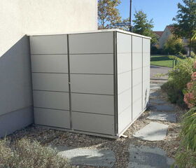 . This image shows a modern, gray outdoor shed, likely made of metal or a similar material. It is situated next to a building and is surrounded by a garden area with stone pathways and some plants. The setting appears to be in a residential area with a street and trees visible in the background.