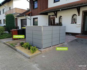 . The image shows a residential area with a structure made of panels outside a house. There are labels indicating the purposes for different sections: "Fahrräder" (bicycles) and "Mülltonnen" (trash bins). Near the structure, there are garbage bins, some small plants, and a paved driveway. The house has a modern aesthetic with white walls and wooden accents.
