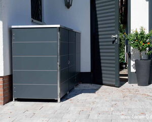 . The image shows a modern garden storage box or cabinet placed next to a house. It is situated on a paved outdoor area. There is a grey door nearby, which is open, and a potted plant with green leaves and small orange flowers next to it. The house exterior is white with a brick base, and there is a wall-mounted light fixture above the cabinet.