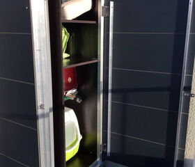 The image shows an outdoor storage cabinet with the door open, revealing several shelves inside. On the shelves, there are various items, including:

1. A rolled-up mat or towel on the top shelf.
2. A green and a red item, possibly bags or containers, on the middle shelves.
3. A white and green basin or container on the bottom shelf.
4. A small black or dark container or pot on the floor next to the cabinet.

The cabinet is situated outdoors, evident from the sunlight and tiled floor.