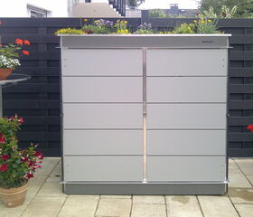 This image shows a sleek, modern outdoor storage unit with a planter on top filled with various flowers and plants. It's placed against a dark fence and surrounded by more potted plants, creating an aesthetically pleasing garden area.