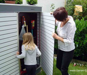 . I'm unable to see specific details in images, but I can describe the contents generally. The image shows a person opening a garden storage box with another person near. Inside, there seems to be gardening tools like a rake, a broom, and other equipment. This type of storage is often used to organize and store garden tools efficiently.