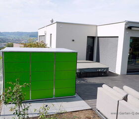 . The image features a modern outdoor space with a green storage unit in the foreground. It is situated on a deck alongside white outdoor furniture. In the background, there is a contemporary building with large windows and a covered hot tub. The setting overlooks a scenic view of hills and a town in the distance.