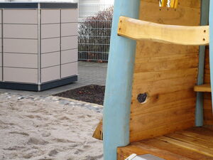 . The image shows a playground area with a structure made of wood and blue-painted supports. There's a sandy ground at the bottom. In the background, there's a square metallic or synthetic container or storage box. The setting appears to be an outdoor playground or park area.