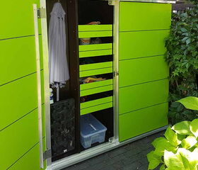 . This image shows a green outdoor storage cabinet with open doors. Inside are several shelves containing various items such as a hanging white garment, a black tray, bottles, and a plastic storage bin. There are also some small items stored on shelves. The setting appears to be a garden or backyard.