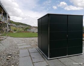 . Ein moderner Schuppen in einem Garten mit gepflastertem Weg und Rasen, umgeben von Landschaft und Häusern.