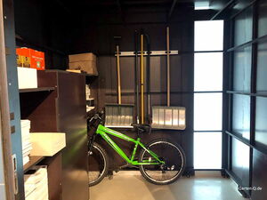 . Ein grünes Fahrrad, Regale mit Kartons und Dokumenten, Wandwerkzeuge wie Schaufeln, in einem dunklen Raum.