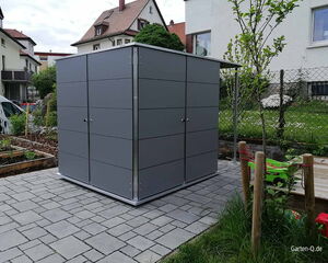 . Ein moderner, grauer Gartenschuppen auf einem gepflasterten Bereich, umgeben von Garten und Häusern.
