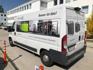 . The image depicts a van with advertising for "Garten[Q]," a company that appears to offer garden-related products. The signage on the van shows images of garden storage solutions, including garden houses, cabinets, and possibly compost bins or similar items. The text on the van is in German and discusses garden houses, garden cabinets, and other storage solutions. The van is parked in an outdoor setting near a building.