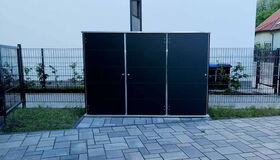 . The image shows a modern outdoor storage shed, likely made of metal or composite materials. It is situated on a paved area with a fence and some greenery in the background. The sleek design suggests it is used for storing garden tools or equipment.