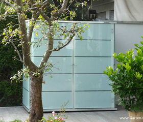 . The image features a modern glass structure with horizontal lines, possibly a shed or storage unit, placed on a patio or outdoor area. There's a tree in the foreground and some greenery, such as bushes or potted plants, surrounding the area.