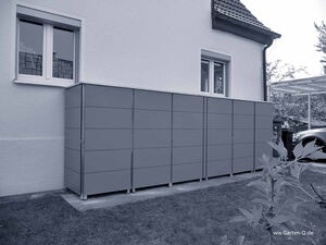 . Grau lackierte Schränke stehen an einer Hauswand mit Fenstern, umgeben von Rasen und Pflanzen.