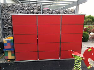 . This image shows a large red structure with horizontal panels, possibly a cabinet or storage unit. Next to it is a transparent container filled with colorful block-like objects. In the foreground, there is a red and green playground spring rocker toy. The background includes a wire mesh filled with rocks and some greenery.