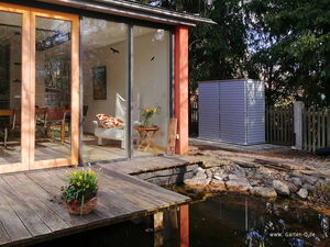 . Holzterrasse mit Blumen, Glaswände zum Innenraum mit Sessel und Tisch, Gartenhütte im Hintergrund.