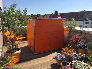 . The image shows a balcony or terrace with an arrangement of various colorful potted plants and flowers. There is also an orange storage unit, as well as an orange folding chair. The background features a view of nearby rooftops on a sunny day.