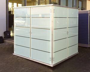 The image shows a cube-like structure made of opaque panels with horizontal black lines, possibly used as a storage unit or booth. It's placed outdoors, near a building entrance. There are bags on the ground nearby, along with a parked chair inside the building. Another similar structure is visible in the background.